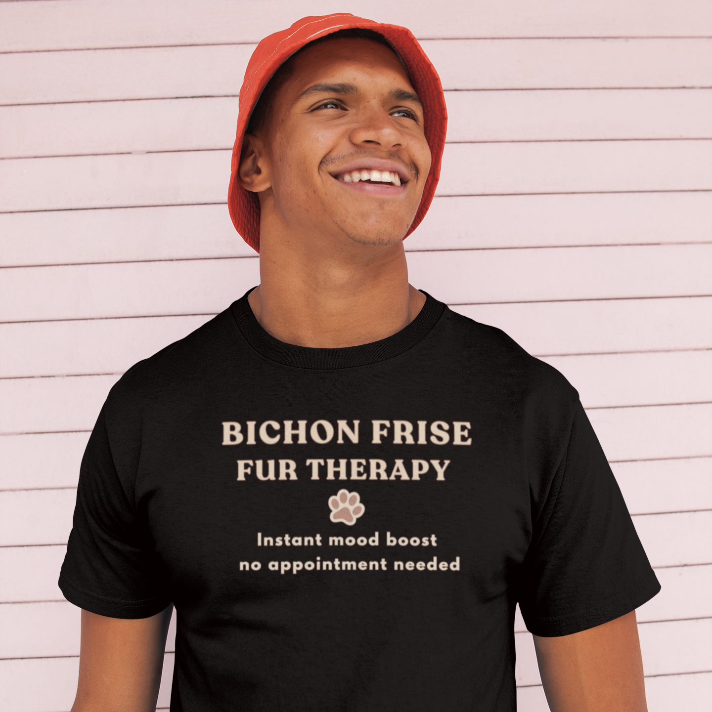 Smiling man wearing a black Bichon Frise Fur Therapy t-shirt with ‘Instant Mood Boost, No Appointment Needed’ slogan, casual outdoor setting with pink wooden wall background.