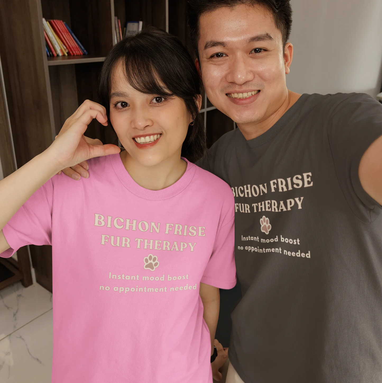 Two smiling young adults wearing Bichon Frise Fur Therapy t-shirts in pink and gray, dog lover apparel, casual style, photographed indoors with a modern background