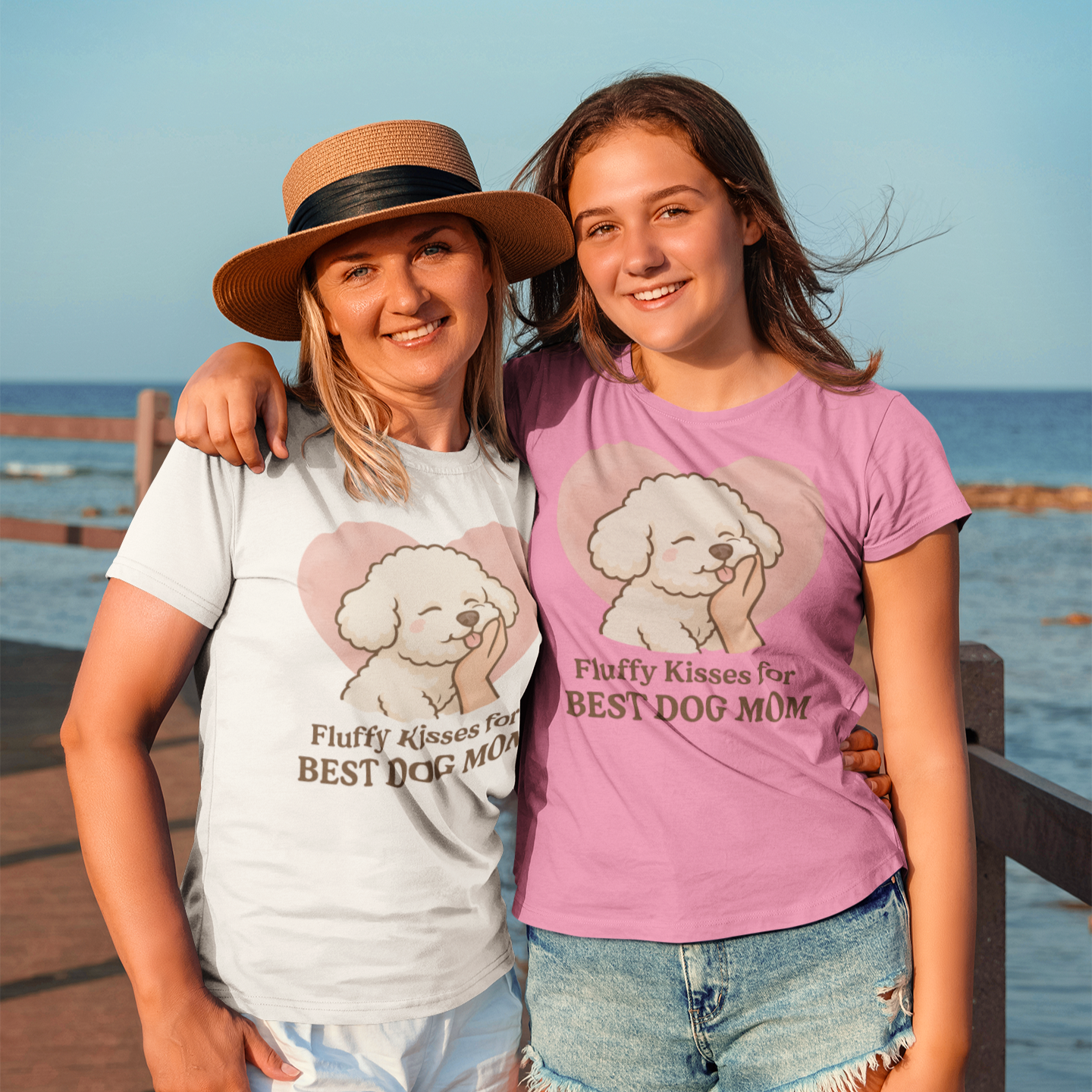 Two smiling women wearing “Fluffy Kisses for the Best Dog Mom” Bichon Frise t-shirts, standing on a sunny boardwalk by the sea – perfect gift for dog moms