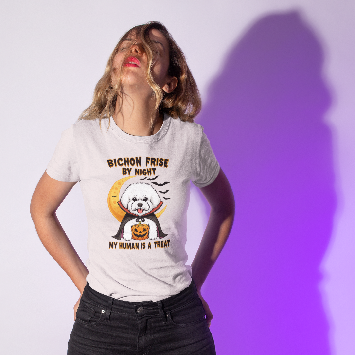 Woman wearing a white T-shirt with a Halloween-themed design of a Bichon Frise dressed as a vampire holding a pumpkin. The shirt text reads “Bichon Frise by Night – My Human Is a Treat”. Purple shadow background