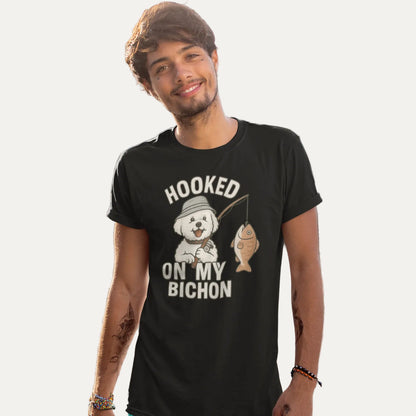 Smiling man wearing a black ‘Hooked on My Bichon’ funny fishing dog dad t-shirt, standing on a sandy beach with boats in the background