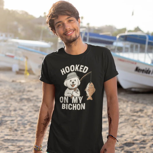 Smiling man wearing a black ‘Hooked on My Bichon’ funny fishing dog dad t-shirt, standing on a sandy beach with boats in the background