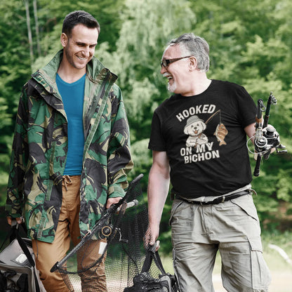 Two men fishing and smiling, one wearing a black ‘Hooked on My Bichon’ funny fishing dog dad t-shirt, outdoors with lush green forest background.