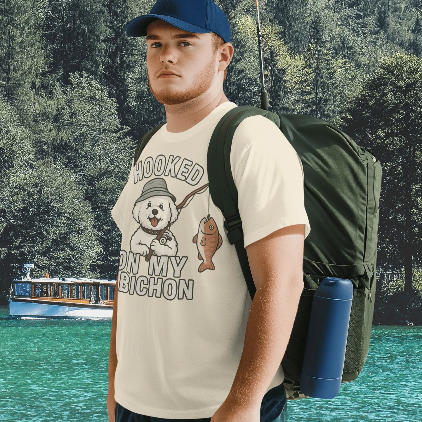 Young man wearing a cream ‘Hooked on My Bichon’ funny fishing dog dad t-shirt with backpack, standing by a lake surrounded by forest
