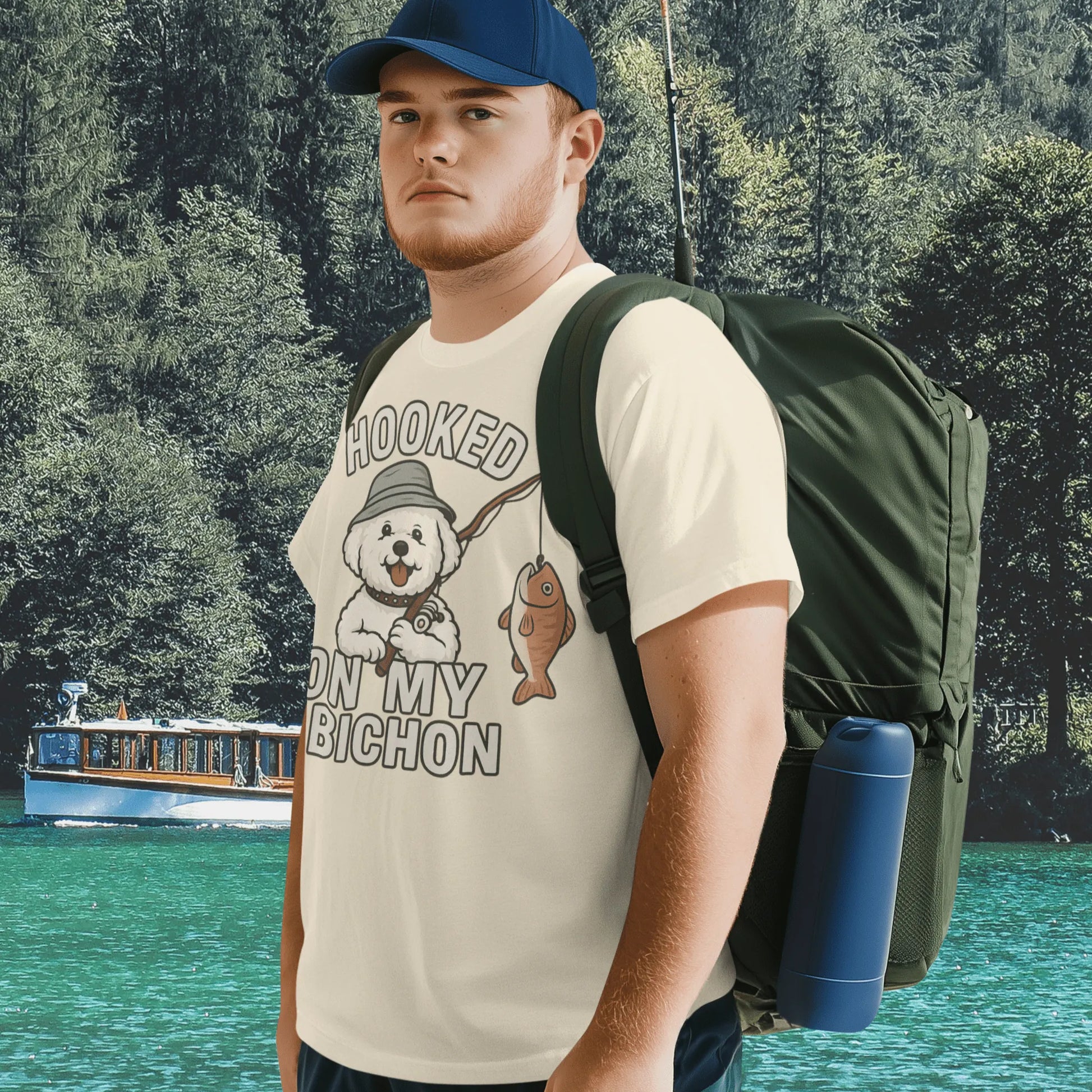 Young man wearing a cream ‘Hooked on My Bichon’ funny fishing dog dad t-shirt with backpack, standing by a lake surrounded by forest