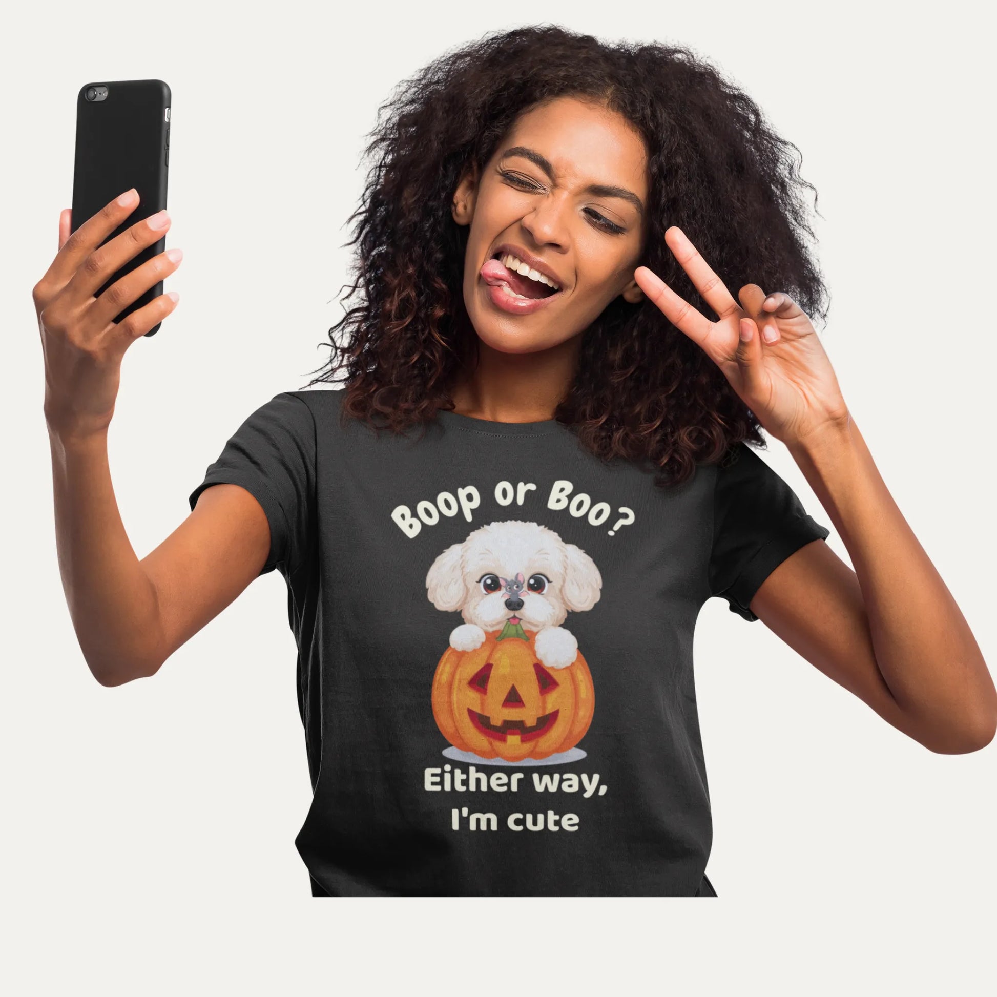 Woman wearing a black Halloween t-shirt with a cute Bichon Frise popping out of a pumpkin and the text “Boop or Boo? Either way, I’m cute” while taking a selfie and making a peace sign – funny dog lover shirt mockup.