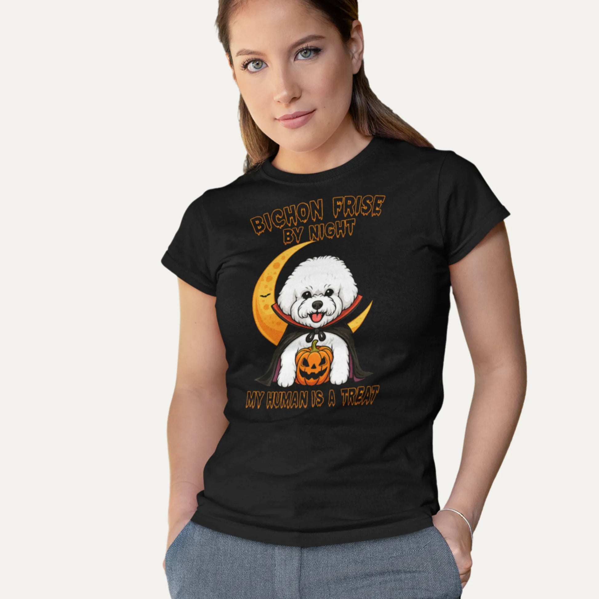 Woman wearing a black T-shirt with a funny Halloween design featuring a Bichon Frise in a vampire costume holding a pumpkin. Text on shirt: “Bichon Frise by Night – My Human is a Treat